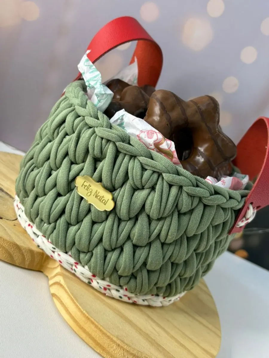 Cesta de Natal em fio de malha verde com alça vermelha, plaquinha Feliz Natal e chocolates dentro